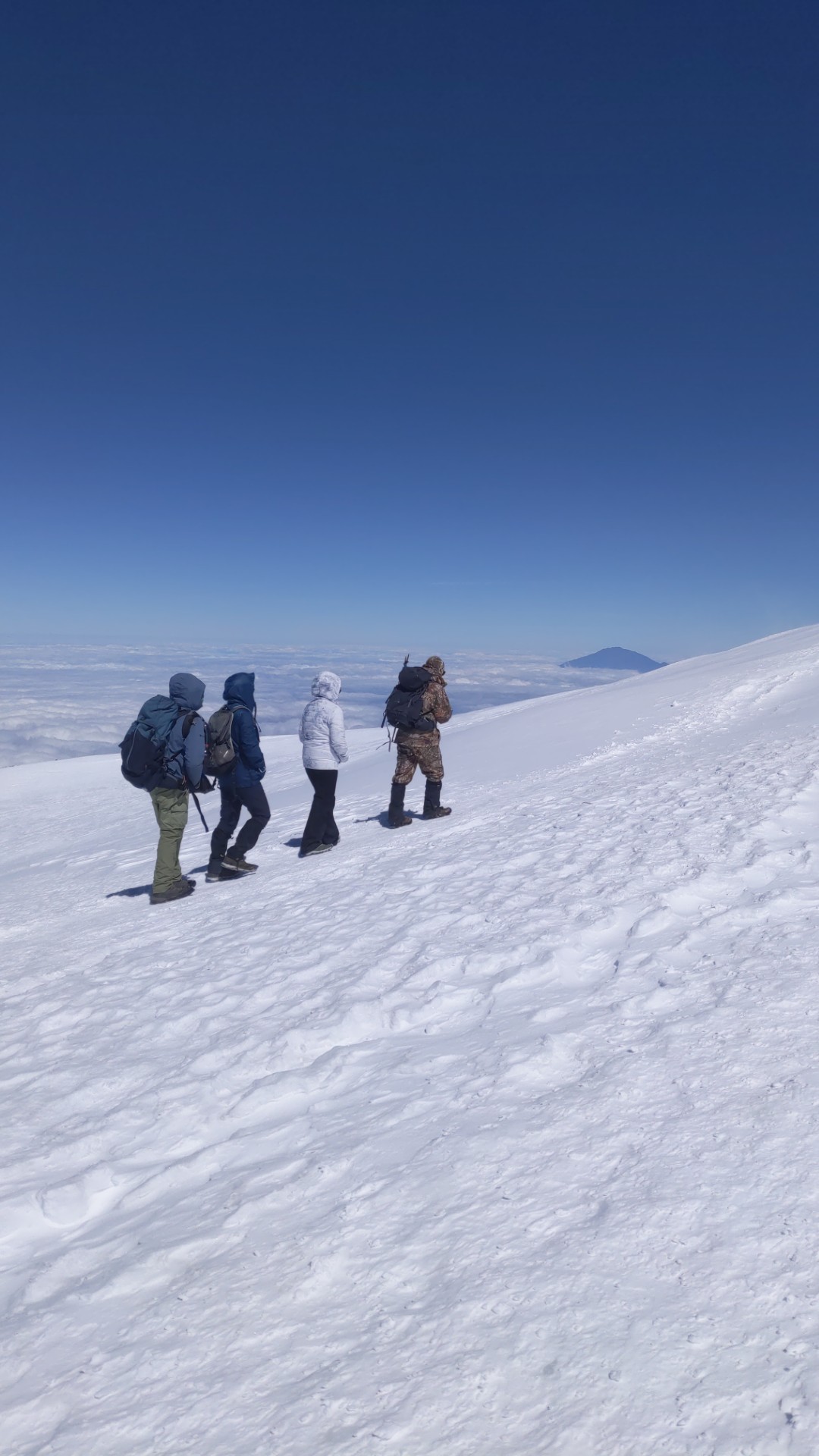 Vier Bergsteiger mit Rucksäcken gehen über einen verschneiten Hang, unter ihnen eine geschlossene Wolkendecke