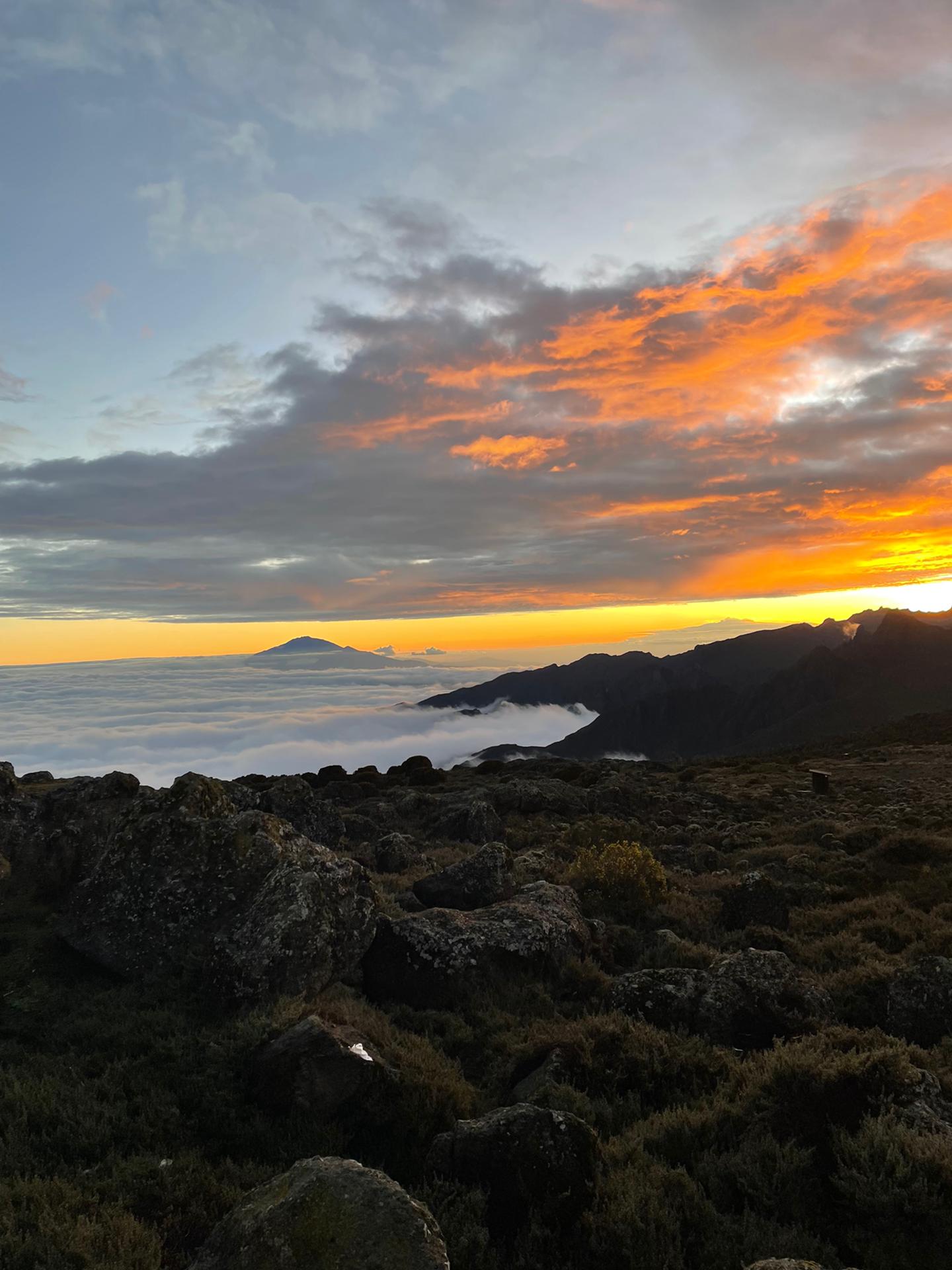 Wolkendecke unter dem Kilimandscharo im warmen Licht der tiefstehenden Sonne, am Horizont die Silhouette des Mount Meru
